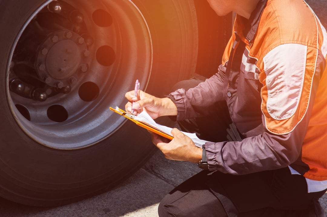 Man with reflective jacket kneeling by truck tire with inspection clipboard.